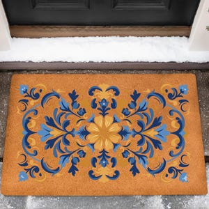 May include: A rectangular doormat with a floral design in shades of blue and yellow on a brown background. The design features symmetrical floral and leaf patterns. The doormat is placed in front of a black door, with snow on the ground.