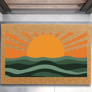 May include: A rectangular doormat with a sunrise design. The sun is a large orange circle with orange rays emanating from it. Below the sun are green and teal waves. The mat is made of a natural fiber material.