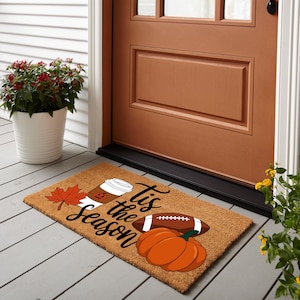 May include: A rectangular coir doormat with a seasonal design. It features a coffee cup with a pumpkin face, a maple leaf, a football, and a pumpkin. The words "Tis the Season" are written in a script font.