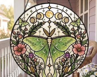 Op de afbeelding: Een ronde glas-in-lood zonnevanger met een grote centrale groene maanspinner, omringd door roze bloemen, paarse klokvormige bloesems en groen blad. Het ontwerp bevat een boog met maanfasen aan de bovenkant en kleine sterren.