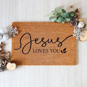 May include: A rectangular doormat made of natural coir fiber. The mat features the words "Jesus LOVES YOU" in a black script font. Decorative elements include silver ornaments, pinecones, and faux greenery.
