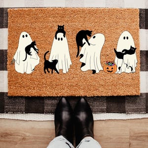 May include: A rectangular doormat with a brown surface features a Halloween design. The design includes four white ghost figures with black cat companions. A jack-o'-lantern with candy sits near one ghost. The mat is placed on a black and white checkered rug.