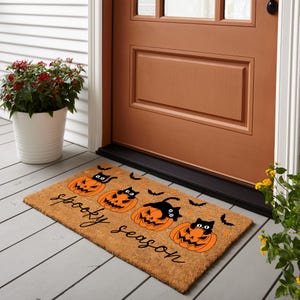 May include: A rectangular doormat with a brown background and the words "spooky season" in black script. The mat features orange jack-o'-lanterns with black cat silhouettes and black bats. The mat is placed in front of a brown door.