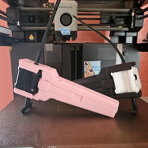 Peut inclure: Deux objets imprimés en 3D, un rose et un noir, sont présentés. L'objet rose est incliné, révélant sa forme. L'objet noir est partiellement visible. Les deux objets ont une conception mécanique complexe. Un fond d'imprimante 3D.