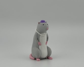 May include: A small, upright figurine of a gray rodent with a white belly, pink paws, and a pink tail. The figure wears a tiny purple hat perched on its head and a white beaded necklace around its neck, featuring dark eyes and a small pink nose.