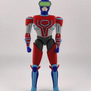 May include: A standing action figure with a blue helmet, green visor, and a red, white, and blue suit. The chest features a red and white geometric emblem. The figure has articulated joints at the shoulders, elbows, and knees, with blue hands and feet.