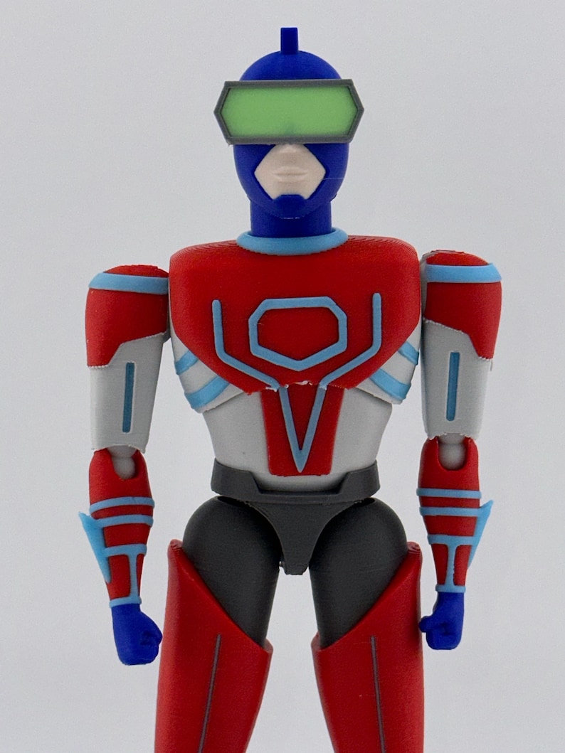 May include: A plastic action figure featuring a blue helmet with a green visor, a red and white chest plate with blue trim, and grey and red limbs. The figure has a stylized humanoid form with articulated joints and a smooth, matte finish.