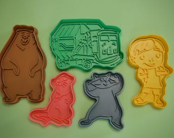 May include: A collection of colorful cookie cutters in the shapes of a bear, a garbage truck, a person, a raccoon, and an otter. The bear is brown, the truck is green, the person is yellow, the raccoon is gray, and the otter is pink.