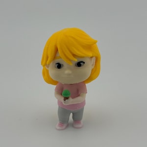 Pode incluir: Pequena estatueta de uma pessoa com cabelo amarelo, vestindo uma camisa rosa, calças cinzentas e sapatos rosa. A figura está segurando um sorvete verde. A estatueta é uma representação de desenho animado.