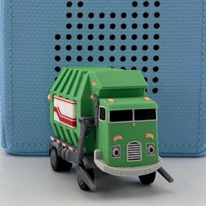 May include: A green toy garbage truck with a white and red design on the side. The truck has a smiling face and is set against a blue background with black dots. The truck has a gray lifting arm.