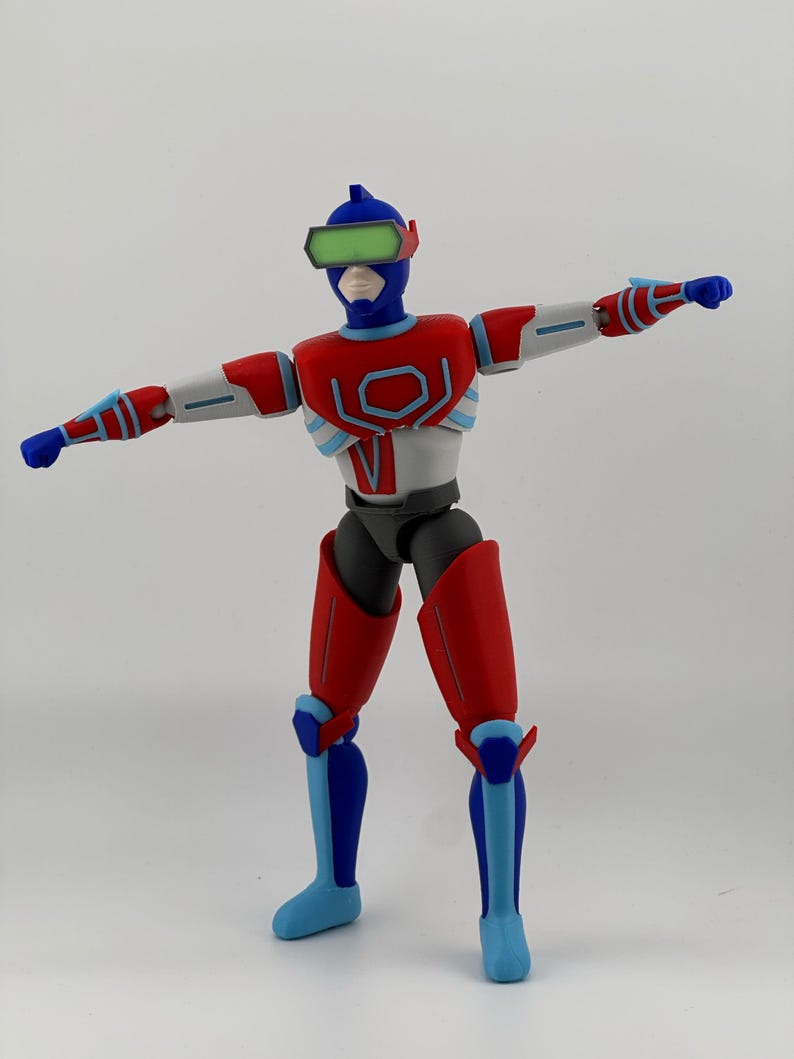May include: A posable action figure featuring a blue, red, and white color scheme. The figure wears a blue helmet with a green visor, a red and white chest plate with a geometric design, and matching red and blue limb armor with blue fists.