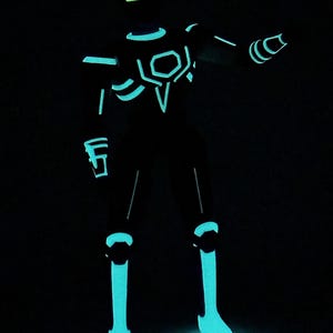 May include: A humanoid action figure standing in a dark environment, featuring glowing cyan and neon green light-up accents across its body. The figure has a rectangular visor, geometric chest plating, and illuminated lines along the limbs.