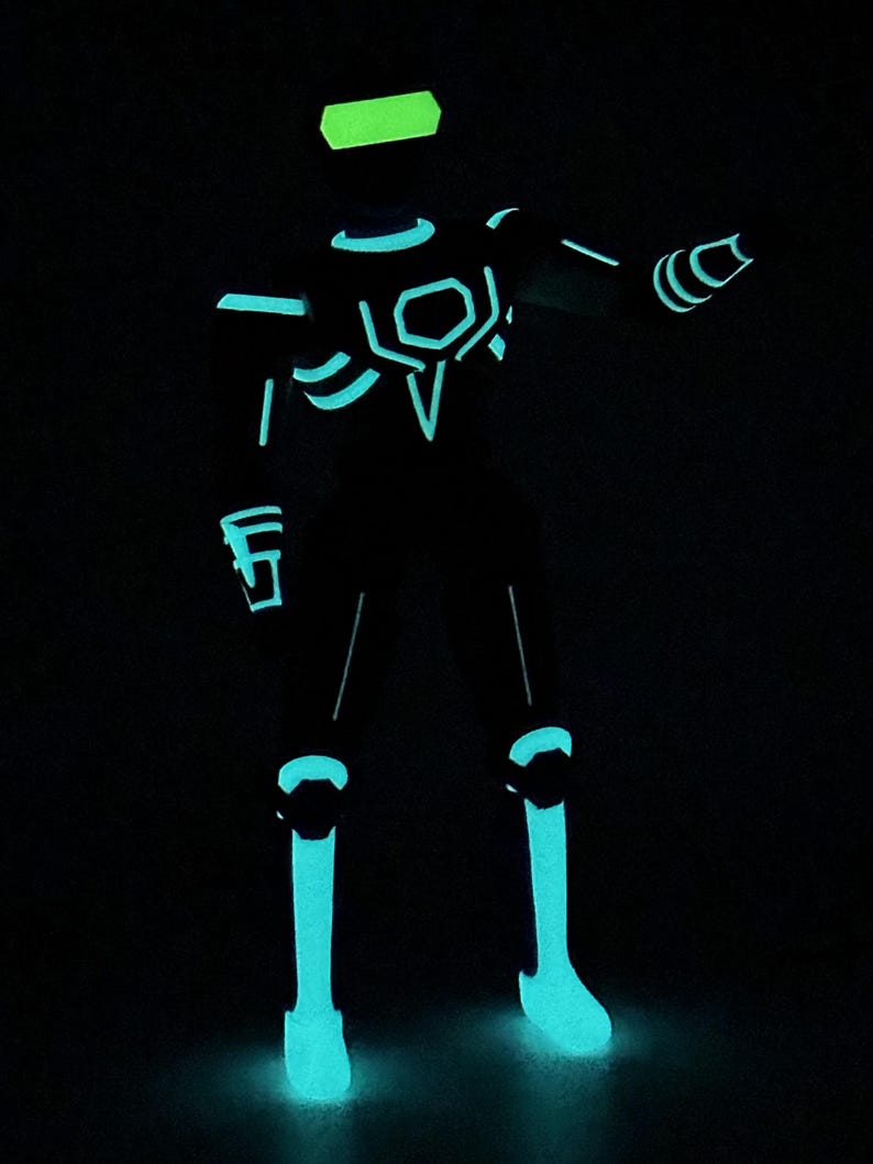 May include: A humanoid action figure standing in a dark environment, featuring glowing cyan and neon green light-up accents across its body. The figure has a rectangular visor, geometric chest plating, and illuminated lines along the limbs.
