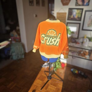 May include: An orange crochet sweater with the word "Crush" in green and yellow lettering on the back. The sweater has a yellow collar and cuffs, and is displayed on a mannequin.