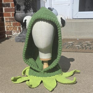 May include: A green crochet hood featuring a frog-inspired design with large white eyes and black pupils on the sides. The hood has a ribbed texture and a collar base with multiple pointed, light green petal-like extensions around the bottom edge.
