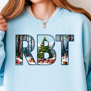 May include: A light blue sweatshirt with the letters "RBT" in a forest scene design. The letters are filled with a winter landscape featuring a Christmas tree, snow, and wrapped gifts. The sweatshirt is a solid light blue color.