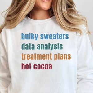 May include: White sweatshirt with the phrases "bulky sweaters," "data analysis," "treatment plans," and "hot cocoa" in blue, green, orange, and red text. The sweatshirt is a crew neck style.
