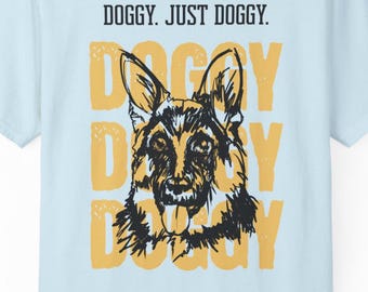 May include: A light blue t-shirt featuring a black ink-style illustration of a German Shepherd dog face with its tongue out. Behind the dog is the word DOGGY printed in large yellow letters three times. The text DOGGY. JUST DOGGY. is above.