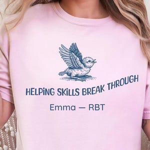 May include: A pale pink t-shirt featuring a blue bird graphic and the text "HELPING SKILLS BREAK THROUGH". The text "Emma - RBT" is below the bird. The word "Blossom" is at the bottom of the image.