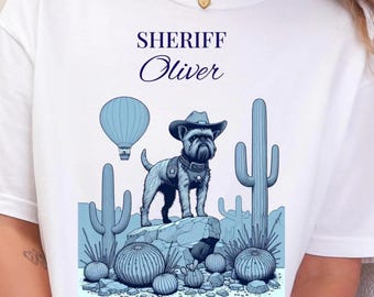 May include: A white t-shirt featuring a blue graphic of a dog wearing a cowboy hat and vest, standing on a rock. The text reads SHERIFF Oliver. The scene includes a hot air balloon and various cacti in shades of blue against a light blue background.