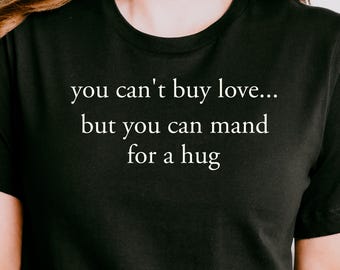 May include: A black short-sleeved t-shirt featuring white text printed on the chest that reads, you can't buy love... but you can mand for a hug. The shirt has a classic crew neckline and is made of a solid black fabric with clean white lettering.