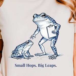 May include: A cream-colored t-shirt featuring a blue ink-style illustration of two frogs. One frog stands holding a clipboard, while the other sits nearby. Below the illustration, the text reads: Small Hops. Big Leaps. The design is centered.