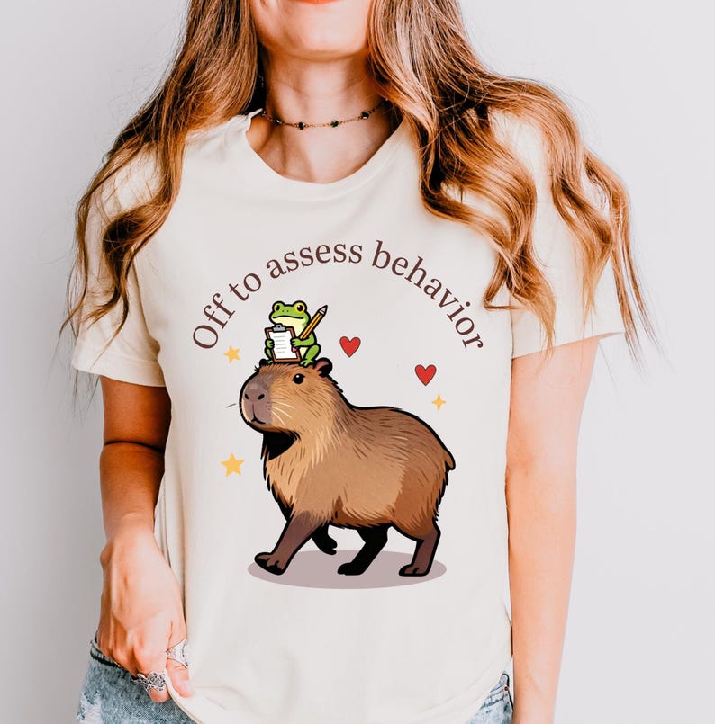 Op de afbeelding: Cr&egrave;mekleurig T-shirt met een afbeelding van een capibara met een kikker op zijn hoofd, die een notitieblok en een potlood vasthoudt. De tekst "Off to assess behavior" staat boven de capibara. Rode harten en gele sterren zijn ook afgebeeld.