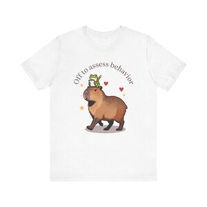 Op de afbeelding: Wit t-shirt met een bruine capibara met een groene kikker op zijn hoofd, die een potlood vasthoudt. De tekst "Off to assess behavior" staat erboven, met rode harten en gele sterren.