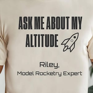 May include: A cream-colored t-shirt featuring the black text "ASK ME ABOUT MY ALTITUDE" above a simple black line-art rocket icon. Below the rocket, the text reads "Riley, Model Rocketry Expert" in a clean, sans-serif black font.