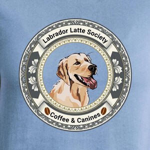 May include: A light blue t-shirt featuring a circular graphic with a yellow Labrador retriever illustration. The text reads Labrador Latte Society and Coffee & Canines. The design includes a decorative border with floral patterns and coffee beans.