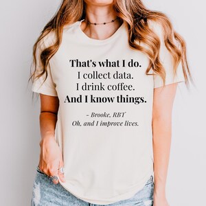 May include: A cream-colored short-sleeve t-shirt featuring black text that reads: That's what I do. I collect data. I drink coffee. And I know things. - Brooke, RBT Oh, and I improve lives. The shirt has a crew neckline and a standard fit.