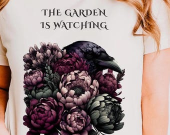 May include: A cream-colored t-shirt featuring a graphic of a black raven emerging from a cluster of dark purple, mauve, and sage green flowers. Above the floral arrangement, the text reads THE GARDEN IS WATCHING in a dark, stylized serif font.