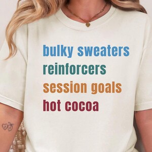 May include: A cream-colored short-sleeve t-shirt featuring four lines of text in different colors. The text reads: bulky sweaters in blue, reinforcers in teal, session goals in orange, and hot cocoa in dark red, all printed in a simple sans-serif font.