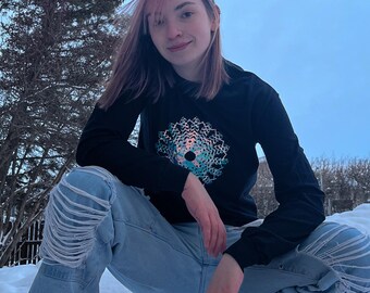 May include: A person wearing a black long-sleeved shirt with a colorful mandala design on the front and blue jeans with distressed details. The person is squatting in the snow.