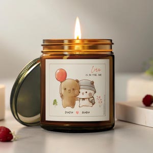 May include: A brown glass candle jar with a white label featuring two cartoon bears holding a red balloon. The label reads "Love is in the air" and "Dudi" and "Bibi" with a heart symbol between the names.