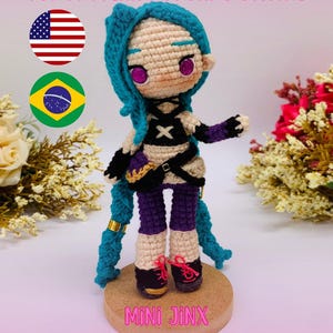 Könnte beinhalten: Gehäkelte Puppe der Figur Jinx aus Arcane, mit blauen Haaren und detailliertem Outfit. Die Puppe steht auf einem kleinen Holzsockel. Der Text lautet "PDF PATTERN / PADRÃO DIGITAL" und "MINI JINX (ARCANE)".