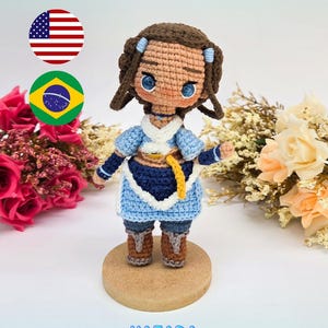 May include: A crochet doll of a character with brown hair, blue eyes, and a blue and white outfit, standing on a circular wooden base. The text "PDF PATTERN / PADRÃO DIGITAL" and "KATARA" appear in blue letters above and below the doll.