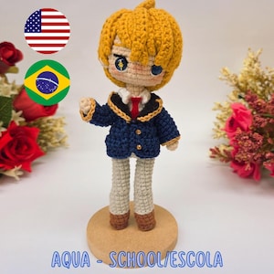 May include: A handmade crochet doll of a character from "Oshi no Ko" wearing a navy blue school uniform with gold accents. The doll has yellow hair and is standing on a wooden base. The text "PDF PATTERN / PADRÃO DIGITAL" and "AQUA - SCHOOL/ESCOLA (OSHI NO KO)" are visible.