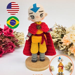 May include: A crochet doll of Aang featuring a blue arrow on the head, wearing a yellow tunic, grey pants, and a red cape. The doll stands on a circular wooden base. The text PDF PATTERN / PADRÃO DIGITAL and AANG are visible in the image.