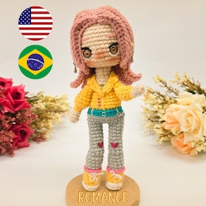Pode incluir: Boneca de crochê com cabelo rosa, jaqueta amarela, calças cinzas com corações e sapatos amarelos. A boneca está em uma base de madeira. O texto na imagem diz "PDF PATTERN / PADRÃO DIGITAL" e "ROMANCE".