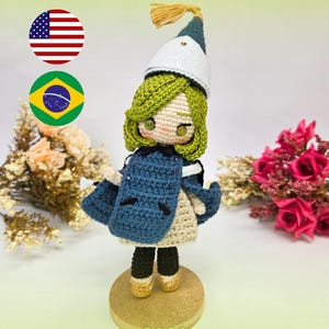 May include: A crochet doll with green hair, wearing a white and blue pointed hat with a tassel, a blue cape, a cream-colored dress, and black pants with tan boots. The doll stands on a circular wooden base, with the text COCO at the bottom.