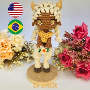 Pode incluir: Boneca de crochê artesanal com pele castanha, usando um top amarelo e calças creme. A boneca tem cabelo loiro encaracolado e pequenos chifres. A boneca está segurando uma pequena flor e está em pé sobre uma base de madeira. O texto na parte superior diz "PDF PATTERN / PADRÃO DIGITAL".