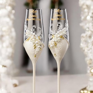 May include: Two champagne flutes with ivory bases and stems. The upper portion of the flutes is clear glass, decorated with white floral designs, pearls, and gold accents. The flutes have personalized text in gold. Elegant wedding or anniversary glassware.