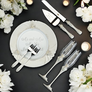 K&ouml;nnte beinhalten: Elegantes Hochzeits-Servier-Set mit Messer und Tortenheber, zwei Sektgl&auml;sern, zwei Gabeln und einem dekorativen Teller. Das Set ist silber-wei&szlig;, mit Strass-Akzenten. Der Teller tr&auml;gt die Aufschrift "Mr and Mrs" und das Datum.