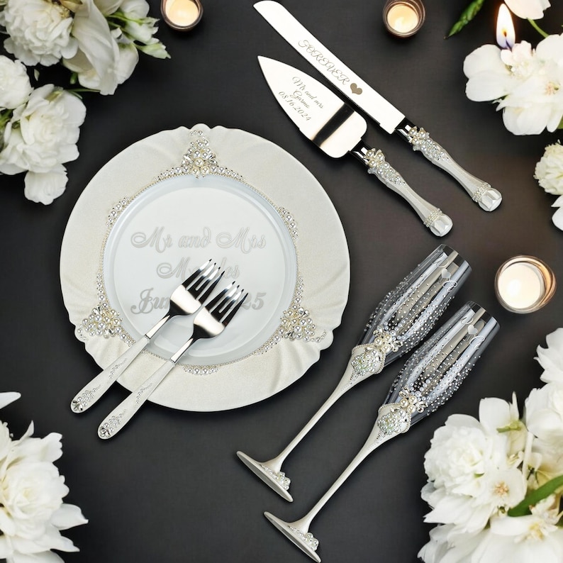 K&ouml;nnte beinhalten: Elegantes Hochzeits-Servier-Set mit Messer und Tortenheber, zwei Sektgl&auml;sern, zwei Gabeln und einem dekorativen Teller. Das Set ist silber-wei&szlig;, mit Strass-Akzenten. Der Teller tr&auml;gt die Aufschrift "Mr and Mrs" und das Datum.