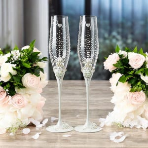 Op de afbeelding: Twee champagneglazen van helder glas met gegraveerde hartmotieven, gedetailleerde witte parelaccenten en verspreide doorzichtige strass-steentjes. De stelen zijn versierd met clusters van parels en kristallen op ronde voetstukken.