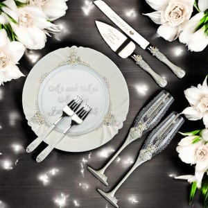 Op de afbeelding: Een trouwset bestaande uit een wit bord met de tekst Mr and Mrs, twee zilveren vorken, een taartmes en taartschep met Forever-gravure en datum, en twee met kristallen versierde champagneglazen met zilveren accenten en strass-steentjes.