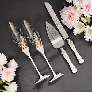 May include: Elegant wedding champagne flutes and cake serving set. The flutes are clear with gold accents and embellished with crystals. The cake knife and server have white handles with crystal and pearl details. The set is personalized with names and a date.