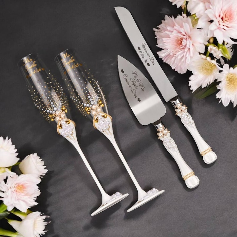 May include: Elegant wedding champagne flutes and cake serving set. The flutes are clear with gold accents and embellished with crystals. The cake knife and server have white handles with crystal and pearl details. The set is personalized with names and a date.