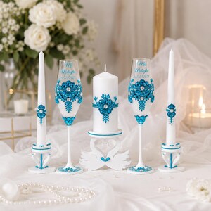 May include: A set of two white taper candles, two champagne flutes, and one pillar candle, all decorated with blue rhinestones and pearls. The flutes feature the text "Mu Malagha 25/12/2025". The pillar candle rests on a white swan-shaped stand.